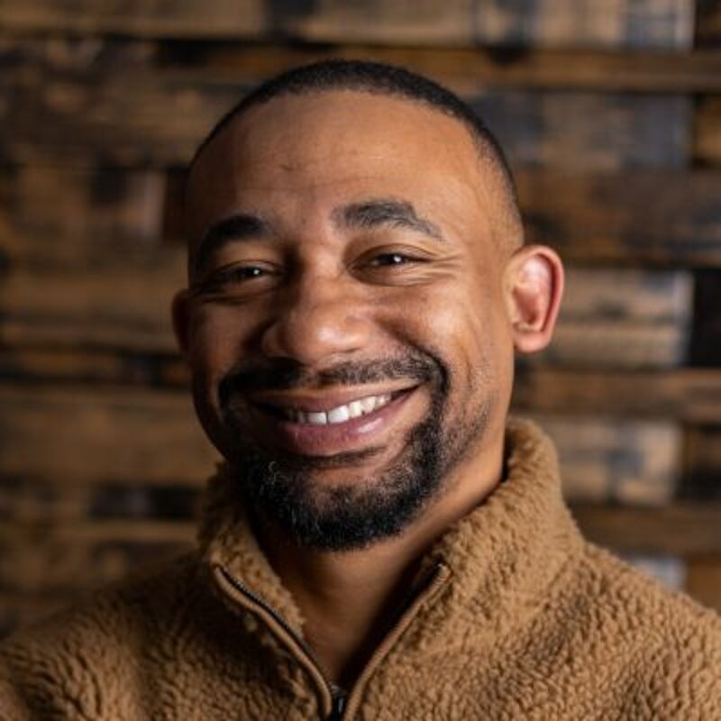 The image shows a close-up of a smiling Black man. He has short, dark hair and a neatly trimmed beard. He is wearing a brown, fuzzy jacket. The background is a wooden wall. The man appears to be in a good mood.